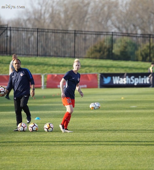 Individuals are engaged in a soccer training session on a grassy field. Some are dribbling soccer balls while a coach oversees the activities. In the background, there is a fence and a sign with the words 'Wash Spirit'.