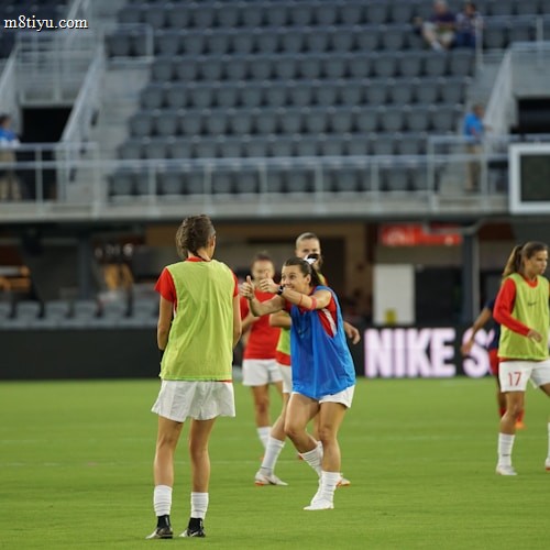 A group of female soccer players are on a field, wearing training bibs over their uniforms. They are practicing with a soccer ball, engaged in a passing drill. The stadium appears empty except for a few spectators and staff. The players are focused and in motion, showing dynamic movement.