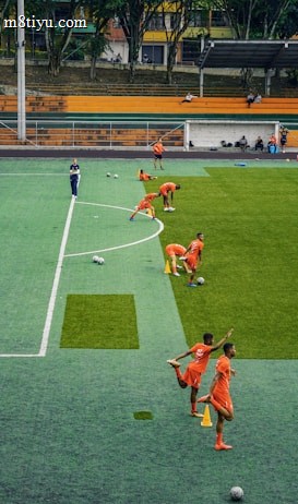 A group of people wearing bright orange sportswear are practicing on a green field. They appear to be athletes, likely soccer players, stretching and preparing for training. There are several soccer balls on the field, and a coach is observing from a distance. The field is surrounded by bleachers with a few onlookers seated, and the area is bordered by trees and residential buildings in the background.