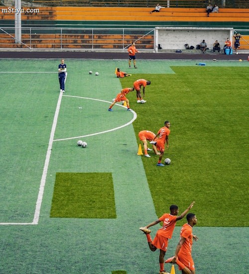 A group of people wearing bright orange sportswear are practicing on a green field. They appear to be athletes, likely soccer players, stretching and preparing for training. There are several soccer balls on the field, and a coach is observing from a distance. The field is surrounded by bleachers with a few onlookers seated, and the area is bordered by trees and residential buildings in the background.