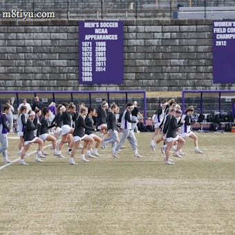 A group of athletes in sports attire are exercising or warming up on a grassy field. They are in a formation, appearing to be mid-stride, and the background shows a series of purple banners with sports achievements listed. The area is surrounded by a stone wall with benches and equipment visible.