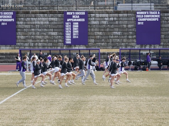 A group of athletes in sports attire are exercising or warming up on a grassy field. They are in a formation, appearing to be mid-stride, and the background shows a series of purple banners with sports achievements listed. The area is surrounded by a stone wall with benches and equipment visible.