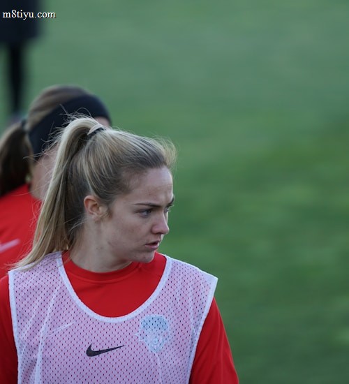 A person with a ponytail wearing a red sports jacket and a white training bib stands on a grassy field. The individual appears to be focused or concentrating, with others in similar attire visible in the background.