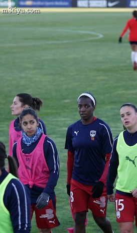A group of female soccer players wearing training vests and sports attire walking on a green soccer field. Some players are wearing pink vests while others are in neon green. They are participating in a training session, with one player slightly in the background. The field is large and appears well-maintained, bordered in the distance by advertising boards.