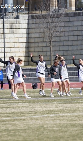 A group of athletes warming up or practicing on a grassy field near a track. They are wearing athletic gear, including shirts and shorts, and appear to be involved in a group exercise or routine. Most have one arm raised, and they are positioned in a line with a large stone wall in the background. A sign on the wall reads 'CAA', indicating a sports conference or event.