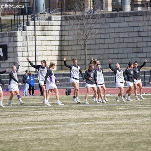 A group of athletes warming up or practicing on a grassy field near a track. They are wearing athletic gear, including shirts and shorts, and appear to be involved in a group exercise or routine. Most have one arm raised, and they are positioned in a line with a large stone wall in the background. A sign on the wall reads 'CAA', indicating a sports conference or event.