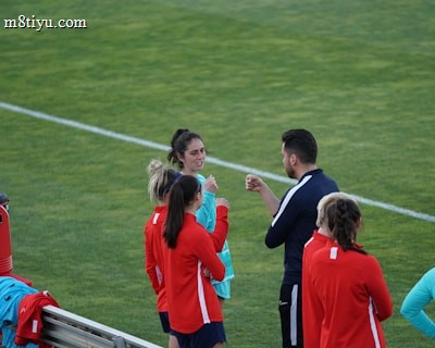 A group of people wearing sports attire are gathered on a grassy field. Some individuals are dressed in red jackets, while others are in turquoise and navy blue. A coach appears to be giving instructions. Sports equipment, such as water bottles and a bench with towels, can be seen nearby.