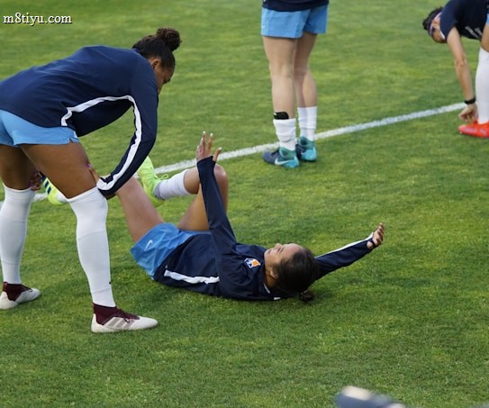 A group of athletes in sportswear are on a green soccer field. Two individuals are interacting closely; one helps the other who is lying on the grass. Nearby, other athletes are stretching.