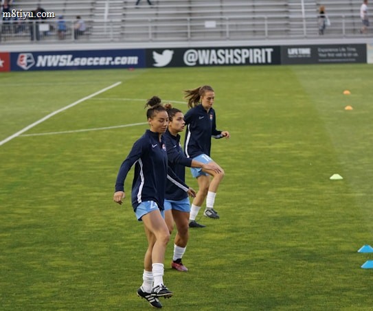Three athletes are warming up on a grassy field, each wearing navy blue jackets and light blue shorts. They are engaged in a drill involving small orange and blue cones placed on the field. Spectators can be seen sitting in the background in a stadium setting, and advertisements are displayed on the side of the field.