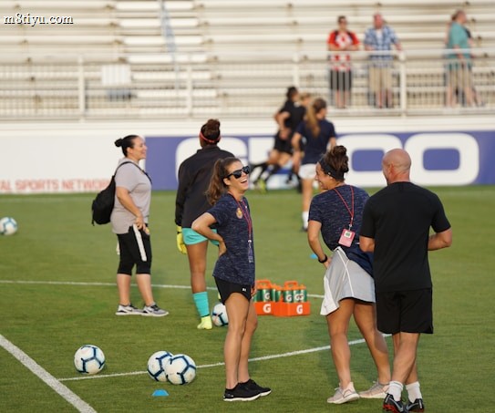 A group of people are gathered on a soccer field, likely during a training session. Some individuals are wearing athletic gear and appear to be coaching or instructing, while others are moving in the background. There are soccer balls scattered on the grass, and orange cones mark certain areas. A nearby bench is visible with some seated spectators.