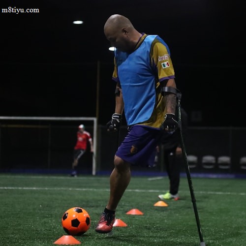 A man wearing a blue training vest is practicing on a football field. He is using crutches and moving a bright orange ball with his foot. Other people in sports attire can be seen in the background, and the field is marked with orange cones.