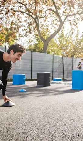 A man in athletic clothing is engaged in an outdoor workout or training session, bent forward while holding a marker or cone. The environment includes other sports equipment such as blue and gray barrels and more cones scattered around. In the background, additional people are seen participating, with trees and a building visible, suggesting a park or recreational area.