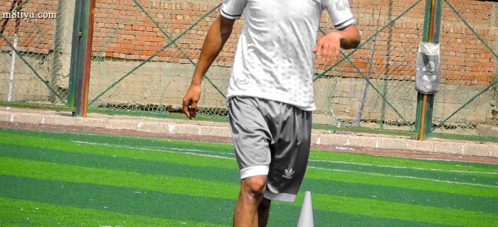 A person in sports clothing is running on a green artificial turf with white and red cones arranged in a pattern. The individual appears to be participating in a sports drill or exercise. The background includes a brick wall and netting consistent with an outdoor sports facility.