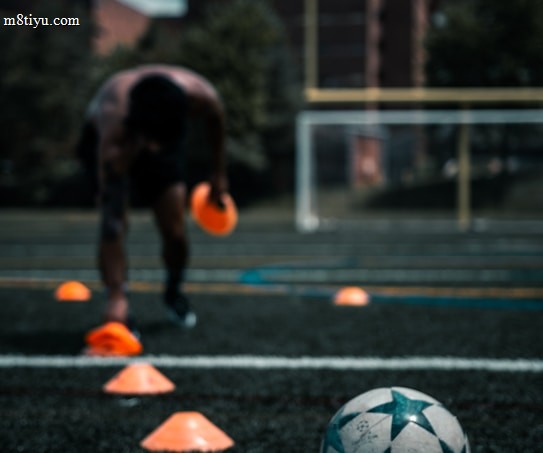 A person is engaged in a soccer training session on an outdoor field. The individual is focused on maneuvering through orange cones arranged on the grass. In the foreground, a soccer ball rests on the ground. There are buildings and trees in the background under a partly cloudy sky.