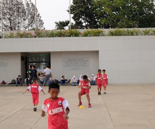 Several children in red sports uniforms are actively participating in a cone drill on a large concrete surface. The background features a modern white building, trees, and a few people sitting or standing near the structure.
